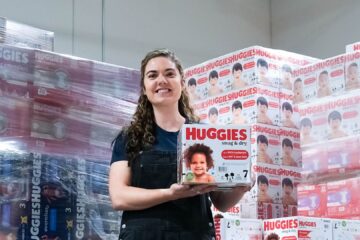 San Diego Food Bank staff member holding up a box of diapers in front of pallets of diapers.