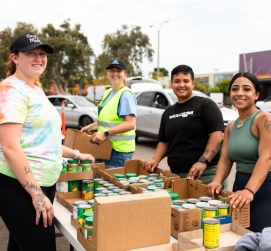 A group of 4 volunteering off-site at a food distribution standing in front of canned food.