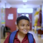 Three elementary-age children wearing backpacks walk down a school hallway.