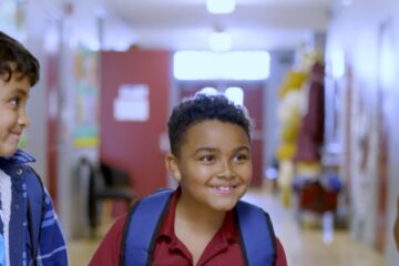 Three elementary-age children wearing backpacks walk down a school hallway.