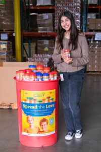 Woman holding a jar of peanut butter beside a red food drive barrel with a “Spread the Love” campaign poster.
