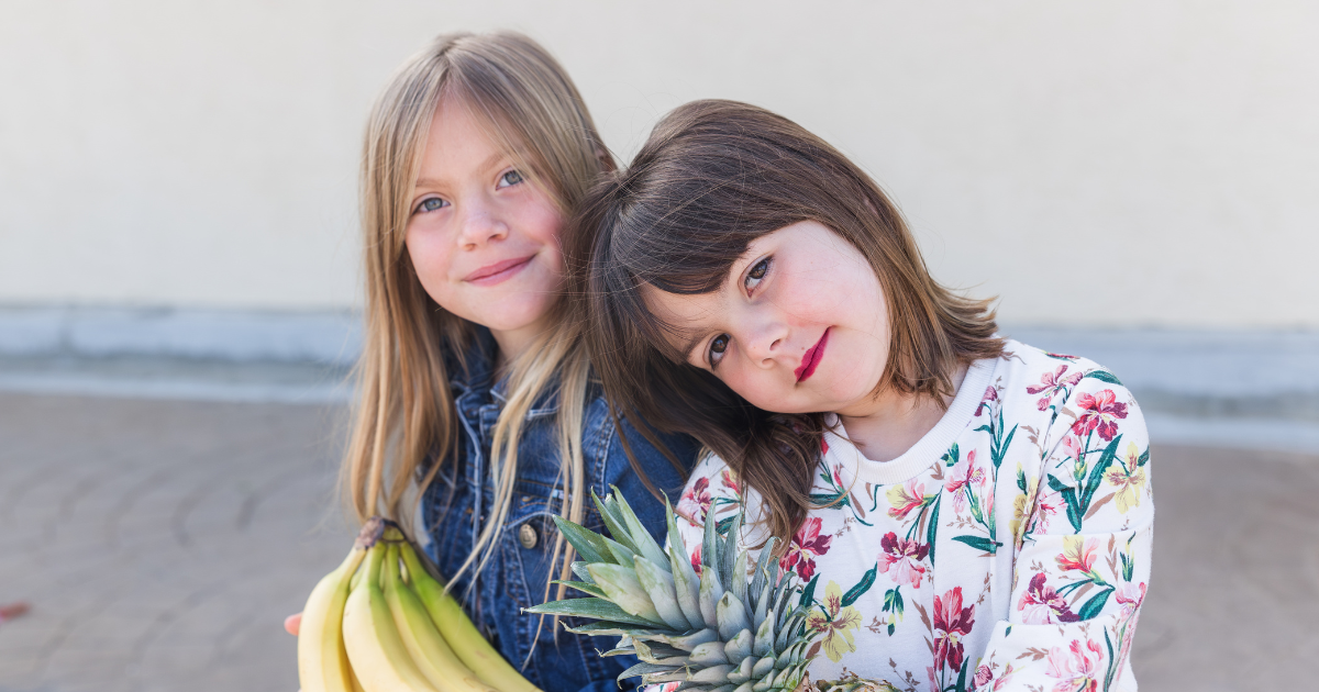 Two sisters sitting next to each other holding fruit.